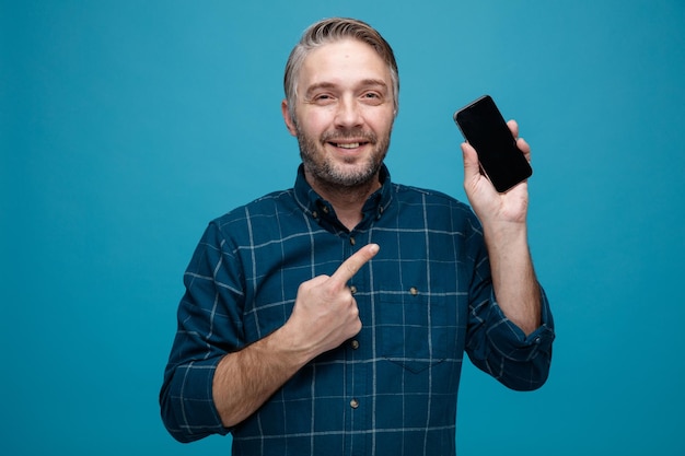 Mann mittleren Alters mit grauen Haaren in dunklem Hemd, der das Smartphone zeigt, das mit dem Zeigefinger darauf zeigt und in die Kamera lächelt, glücklich und positiv über blauem Hintergrund steht