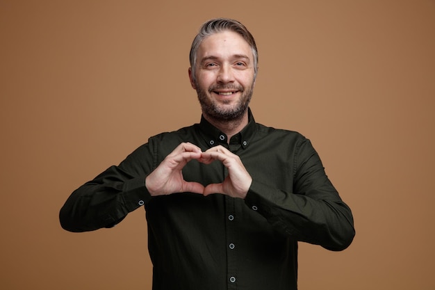 Kostenloses Foto mann mittleren alters mit grauen haaren in dunklem hemd, der glücklich und positiv in die kamera blickt und mit den händen über der brust lächelnd über braunem hintergrund steht