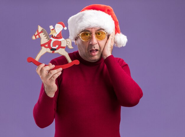 Kostenloses Foto mann mittleren alters mit weihnachtsmütze in gelben gläsern zeigt weihnachtsspielzeug verwirrt über lila wand stehend