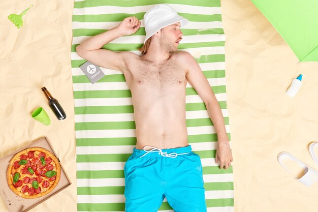 Mann schläft am Strand schnorchelt und sieht süße Träume in der Sonne liegen trägt Sonnenhut-Shorts, umgeben von Pizzabier-Sonnencreme und Hausschuhen, die im Sommer im Urlaub sind