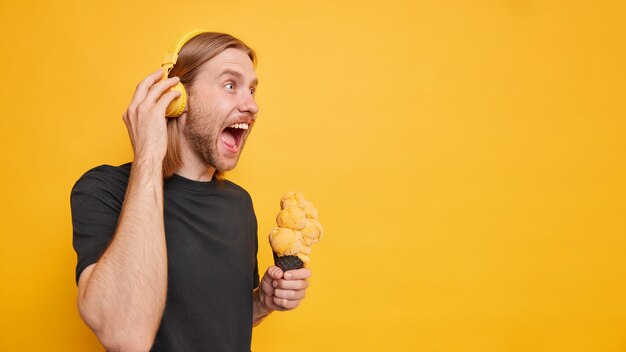 Mann schreit laut öffnet den Mund schreit vor Wut hat roten Bart und Haare hält leckeres Eis in einem lässigen schwarzen T-Shirt gekleidet hört Musik über Kopfhörer isoliert auf gelber Wand