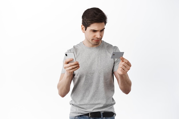 Mann sieht verwirrt aus und plastikkreditkarte, macht online-zahlung, überweist geld in app-smartphone, steht mit telefon vor weißem hintergrund