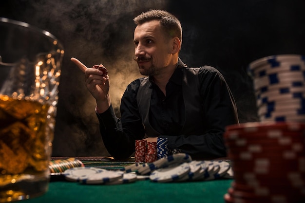 Mann spielt Poker mit einer Zigarre und einem Whiskey. Ein Mann, der mit dickem Zigarettenrauch alle Chips auf dem Tisch gewinnt. Das Konzept des Sieges. Glas mit Whiskey und Stapel Chips im Vordergrund