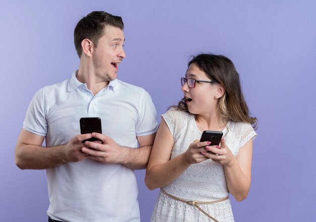 Mann und Frau des jungen Paares, die Smartphones halten, die einander überrascht und glücklich betrachten, über blauer Wand stehend