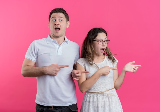 Mann und Frau des jungen Paares in der Freizeitkleidung, die mit den Fingern zur Seite zeigt, überrascht und erstaunt, über rosa Wand stehend