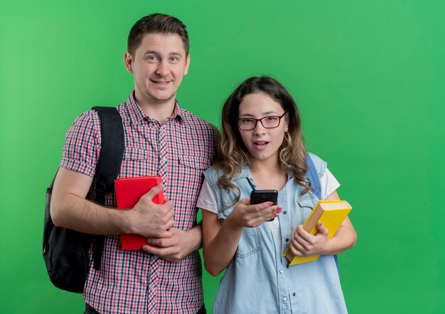 Mann und Frau des jungen Paares in der Freizeitkleidung mit Rucksäcken, die Bücher lächelnd stehen über grüner Wand halten