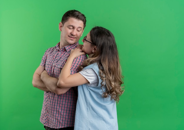 Mann und Frau des jungen Paares in der glücklichen Frau der Freizeitkleidung, die ihren geliebten lächelnden Mann umarmt, der über grüner Wand steht