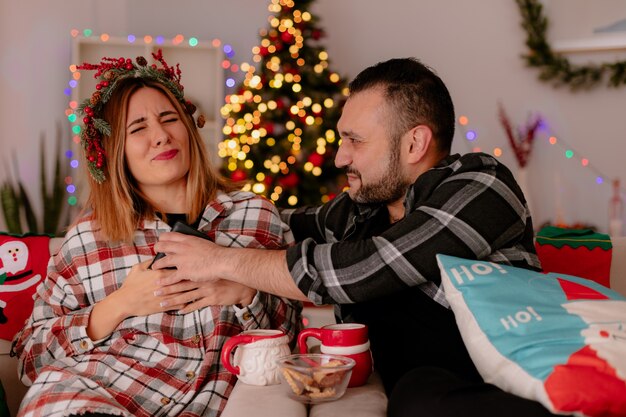 Mann und Frau des jungen Paares mit Smartphone, das auf einer Couch mit Tassen des Teestreit-dekorierten Zimmers mit Weihnachtsbaum im Hintergrund sitzt