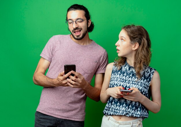 Mann und Frau des jungen Paares mit Smartphones Frau überrascht und verwirrt, ihren Freund über grünem Hintergrund betrachtend