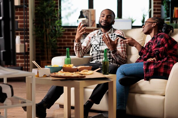 Mann und Frau genießen Fast-Food-Lieferung und Fernsehen, wechseln die Kanäle, um Filme im Fernsehen zu finden. Essen zum Mitnehmen und Bier aus der Flasche trinken, Freizeitbeschäftigung.