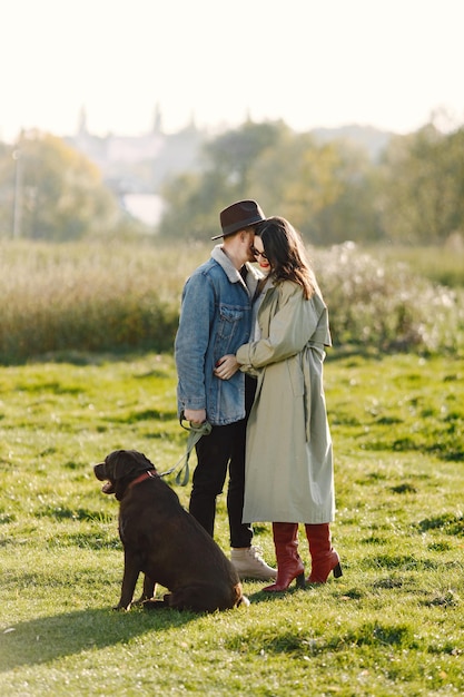 Mann und Frau in modischer Kleidung, die mit ihrem Labrador auf einer Natur ruht. Mann mit Jacke und schwarzem Hut und Frauenrock und roten Stiefeln