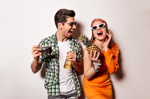 Mann und Frau in stylischen Outfits und Sonnenbrillen lachen aufrichtig und posieren mit Retro-Kamera Ananas und Flasche Bier auf weißem Hintergrund