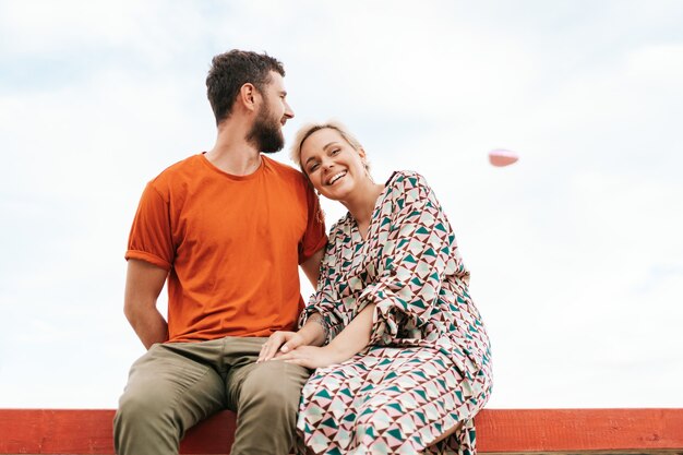 Mann und Frau sitzen glücklich auf einem Holzbrett, das zu einem fliegenden rosa Herzballon im Himmel schaut