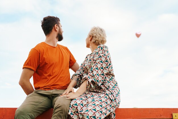 Mann und Frau sitzen glücklich auf einem Holzbrett, das zu einem fliegenden rosa Herzballon im Himmel schaut
