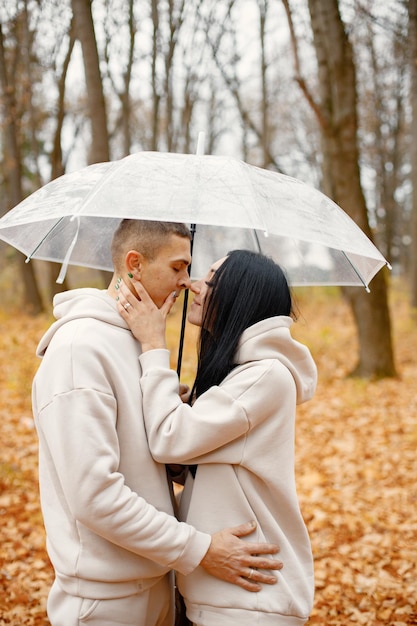 Mann und Frau stehen im herbstlichen Wald und küssen sich unter transparentem Regenschirm Brunette Mann und Frau verlieben sich Familie in beige sportlichen Kostümen