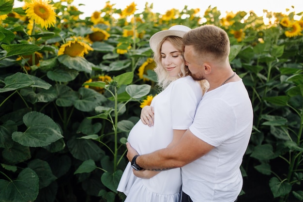 Mann und schwangere Frau umarmen sich zart stehend auf dem Feld mit hohen Sonnenblumen um sie herum