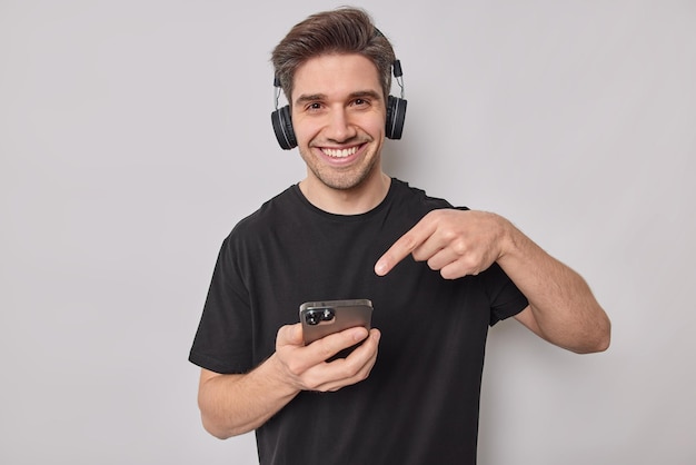 Mann zeigt auf Smartphone-Display trägt drahtlose Kopfhörer an den Ohren Kleider in lässigem schwarzem T-Shirt isoliert auf Weiß zeigt, wie schnell seine neuen Geräte funktionieren.