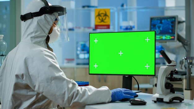 Mannforscher im Overall mit Blick auf den Chroma-Key-Computer in einem modern ausgestatteten Labor. Team von Mikrobiologen, die Impfstoffforschung betreiben und auf Gerät mit grünem Bildschirm schreiben, isoliert, Mockup-Display.