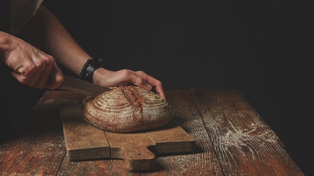 Mannhände schneiden ein frisches rundes Brot auf einem Schneidebrett auf einem hölzernen Hintergrund