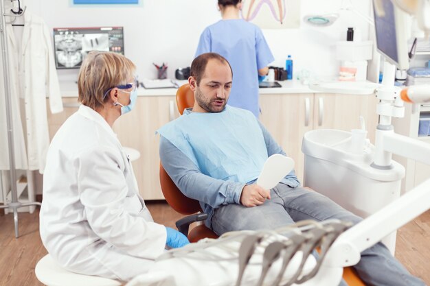 Mannpatient, der in den Spiegel schaut, nachdem das medizinische stomatologische Team die Zahnoperation während des stomatologischen Verfahrens abgeschlossen hat