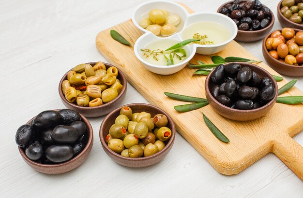 Marinierte Oliven und Olivenöl mit Olivenblättern in Schalen und Schneidebrett auf weißem Holz, Blickwinkel.
