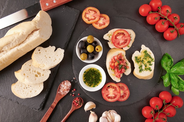 Mediterrane Snacks. Oliven, Öl, Kräuter und geschnittenes Ciabatta auf einem Holzbrett auf schwarzem Schiefersteinbrett über dunkler Oberfläche, saftige Tomaten auf frischem Brot, Pesto als Belag. Draufsicht. Flach liegen