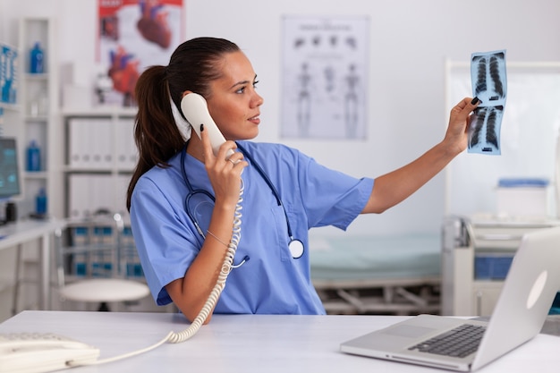 Medizinische Krankenschwester, die Patientenradiographie im Krankenhausbüro hält, während sie mit dem Arzt am Telefon spricht...