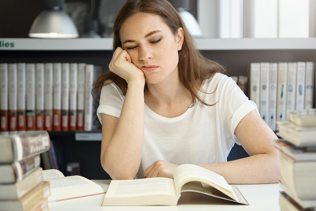 Medizinische Universität kaukasische Studentin, die an der Bibliothek studiert, schöne College-Frau, die schläft, während sie vor einem offenen Buch sitzt, das ihr Kinn auf einer Hand ruht und erschöpft aussieht.