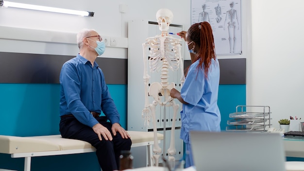 Medizinischer Assistent, der alten Patienten bei der Konsultation während der Covid-19-Pandemie menschliche Skelettknochen zeigt. Krankenschwester und älterer Mann analysieren die Anatomie des Rückenmarks, um eine orthopädische Diagnose zu finden.