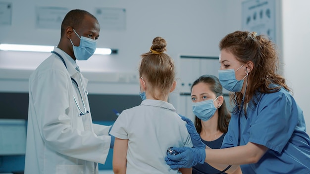 Medizinischer Assistent, der kleines Kind mit Stethoskop im Büro berät. Krankenschwester in uniform, die puls und herzschlag überprüft und während der covid-19-pandemie eine gesundheitsdiagnose findet.