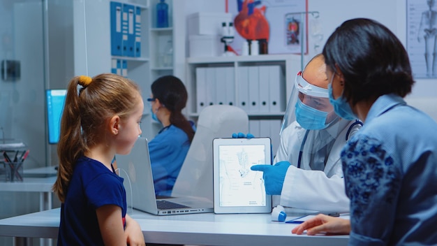 Medizinischer Spezialist, der Skelett mit Tablet auf dem Schreibtisch in der Arztpraxis präsentiert. Kinderarzt mit Schutzmaske, der Gesundheitsdienste, Beratungen und Behandlungen während Covid-19 anbietet