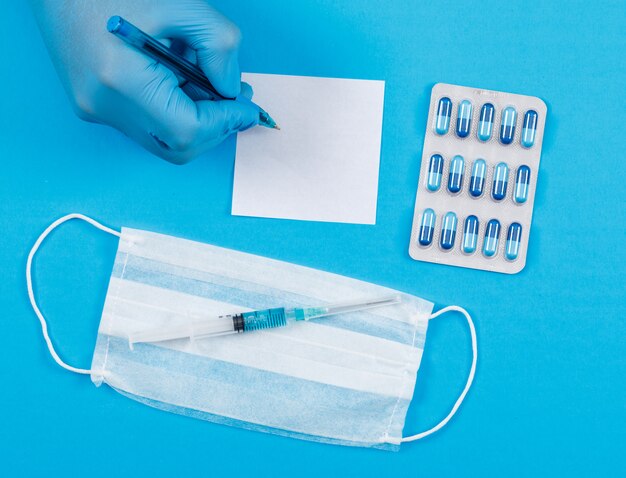Medizinisches Konzept mit Haftnotiz, Packung Kapseln, Gesichtsmaske, Injektion, Haftnotiz auf blauem Schreibtisch-Hintergrund. Hand hält Stift.