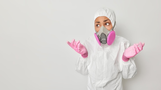 Medizinisches Personal und vorbeugendes Ausrüstungskonzept Verwirrte, zögerliche Frau trägt einen chemischen Schutzanzug mit Kapuzenhandschuhen und Atemschutzmaske gegen Coronavirus-Posen vor grauem Hintergrund, Leerzeichen