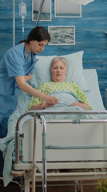 Medizinisches Team, das eine Untersuchung für kranke Patienten im Krankenhausbett in einem Pflegeheim durchführt. Arzt benutzt digitales Tablet, während Krankenschwester einer Rentnerin zur Untersuchung Oximeter zur Hand legt.