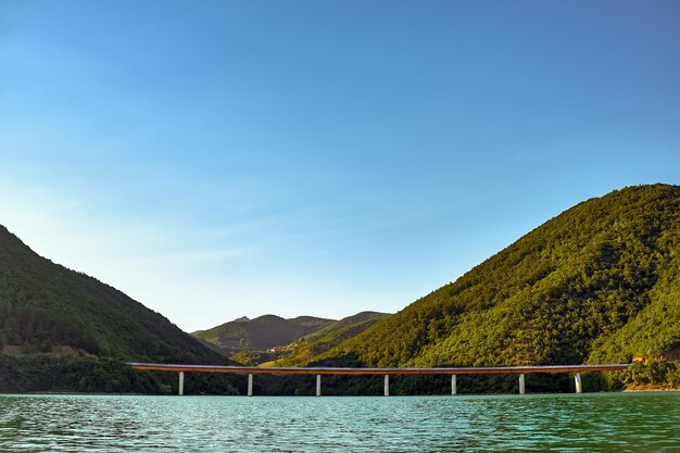 Meer mit einer Betonbrücke, umgeben von Hügeln, die unter dem Sonnenlicht mit Wäldern bedeckt sind