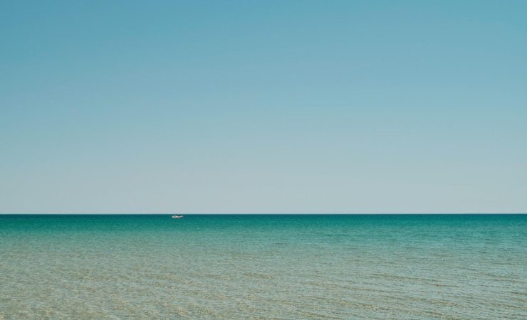 Kostenlose Foto meerespanorama im flachen wasser mit sonnenreflexion im wasser weites offenes meer mit klarer himmelswelle und ruhigem meer mit schönem sonnenlicht rotes boot ist in der ferne sichtbar wallpaper oder banner-idee