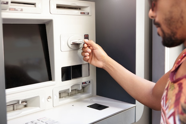 Mensch und Technik. Dunkelhäutiger Mann mit Geldautomat. Die Hand des schwarzen Kerls, die Plastikbankkarte in Geldautomaten oder Geldautomaten einführt