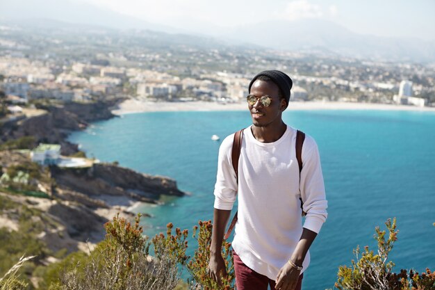 Menschen, aktiver Lebensstil, Reisen, Abenteuer und Tourismuskonzept. Hübscher trendiger aussehender afroamerikanischer Tourist mit Rucksack, der Urlaub im Ausland verbringt