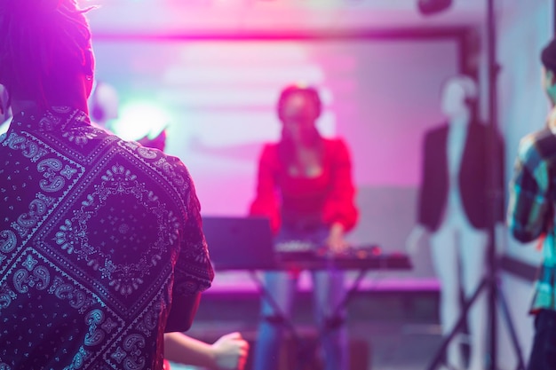 Menschen beobachten ein Live-Konzert elektronischer Musik auf der Bühne mit Scheinwerfern in einem Nachtclub. Ein afroamerikanischer Musiker spielt mit einem DJ-Controller in einem Nachtclub auf einem verschwommenen Hintergrund