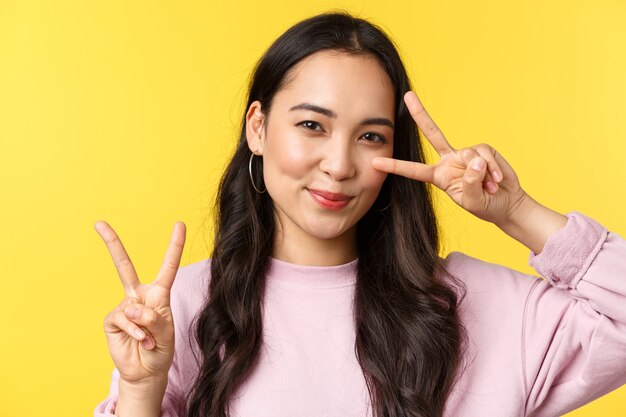 Menschen Emotionen, Lifestyle-Freizeit- und Schönheitskonzept. Kawaii hübsches japanisches Mädchen, das Friedenszeichen zeigt und nett lächelt und über gelbem Hintergrundwerbungsprodukt steht.