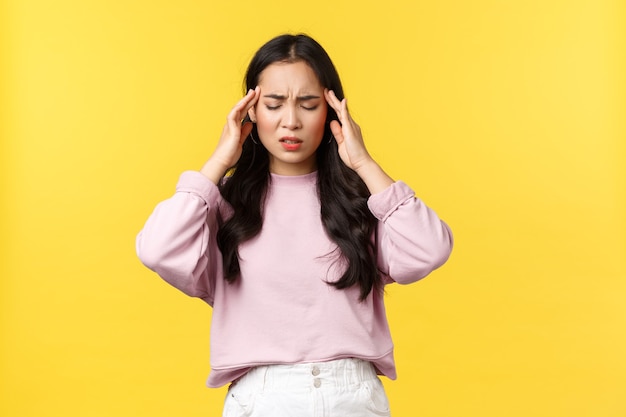 Menschen Emotionen, Lifestyle und Modekonzept. Beunruhigte und erschöpfte asiatische Frau schließt die Augen und berührt die Schläfen, hat Migräne, Kopfschmerzen oder einen schwindligen, gelben Hintergrund.