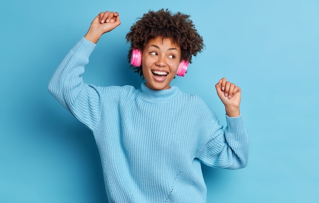 Menschen Entspannung Aktivität Glück Konzept. Zufriedene dunkelhäutige Frau mit Afro-Haar bewegt sich zur Musik trägt drahtlose Stereo-Kopfhörer und lächelt fröhlich, breit gekleidet in einem lässigen Pullover.