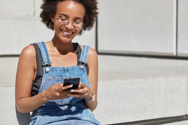Menschen-, Erholungs- und Technologiekonzept. Entspannte sorglose schwarze Frau hält Handy in Händen, tippt SMS an Freund, hat glücklichen Gesichtsausdruck, freien Platz für Ihre Textinformationen