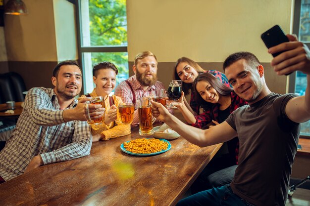 Menschen, Freizeit, Freundschaft und Kommunikationskonzept. glückliche Freunde trinken Bier, reden und klirren Gläser an der Bar oder im Pub und machen Selfie-Foto per Handy.