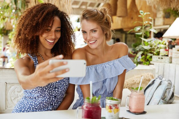 Menschen, Freizeit und Erholungszeit. Fröhliche Afroamerikanerin und ihre beste Freundin verbringen ihre Freizeit im Café, machen Selfies auf dem Handy, trinken Smoothie. Multiethnisches Beziehungskonzept