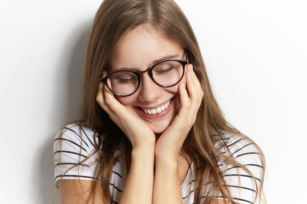 Menschen, Freude, Jugend und Glückskonzept. Schließen Sie herauf Bild des charmanten schüchternen Teenager-Mädchens in der stilvollen transparenten Brille, die schüchtern nach unten schaut und breit lächelt, Gesicht berührt, verlegen ist