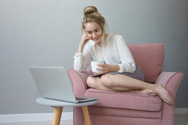 Menschen, Geräte und modernes Technologiekonzept. Schöne fröhliche junge kaukasische Frau, die sich nach der Arbeit zu Hause entspannt, Tee trinkt, lässig im Sessel sitzt und Komödie auf Laptop-PC sieht