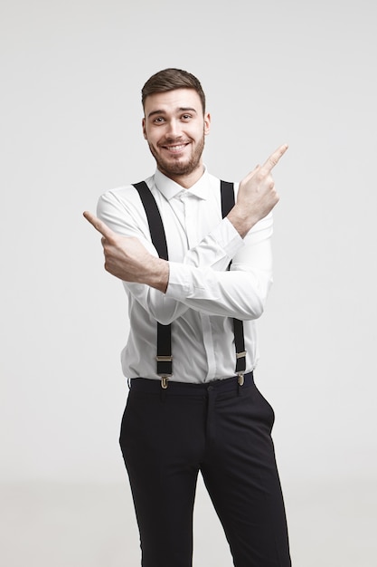Menschen-, Geschäfts-, Job-, Erfolgs- und Karrierekonzept. Bild des emotionalen positiven jungen kaukasischen Geschäftsmannes mit getrimmtem Bart, der aufgeregt lächelt und Zeigefinger in entgegengesetzte Richtungen zeigt