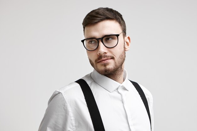 Menschen-, Geschäfts-, Stil-, Mode- und Modellierungskonzept. Attraktives bärtiges junges europäisches männliches Modell in stilvoller Brille und elegantem weißem Hemd mit Hosenträgern, die im Studio für Werbung aufwerfen