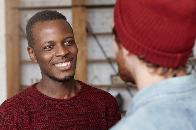 Menschen, interracial Freundschaft und Kommunikationskonzept. Hübscher fröhlicher junger afroamerikanischer Mann in gemütlichem Pullover, der glücklich lächelt und sich darüber freut, dass sein kaukasischer Freund geheiratet hat